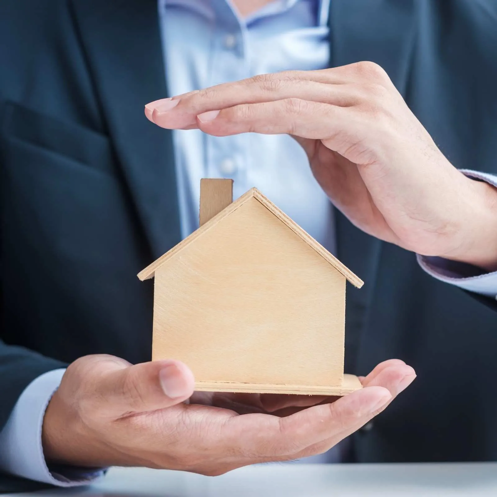 Hands holding a small wooden house model with another hand sheltering it from above